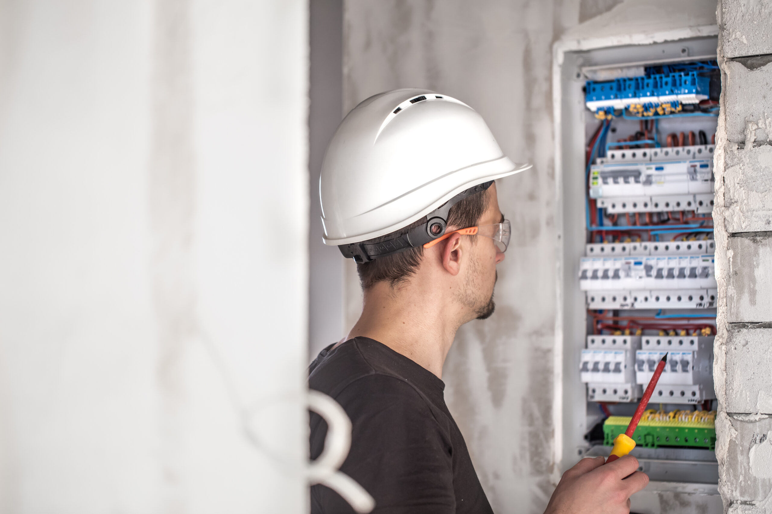 Electrician wearing a white hard hat inspecting a residential circuit breaker panel with a screwdriver